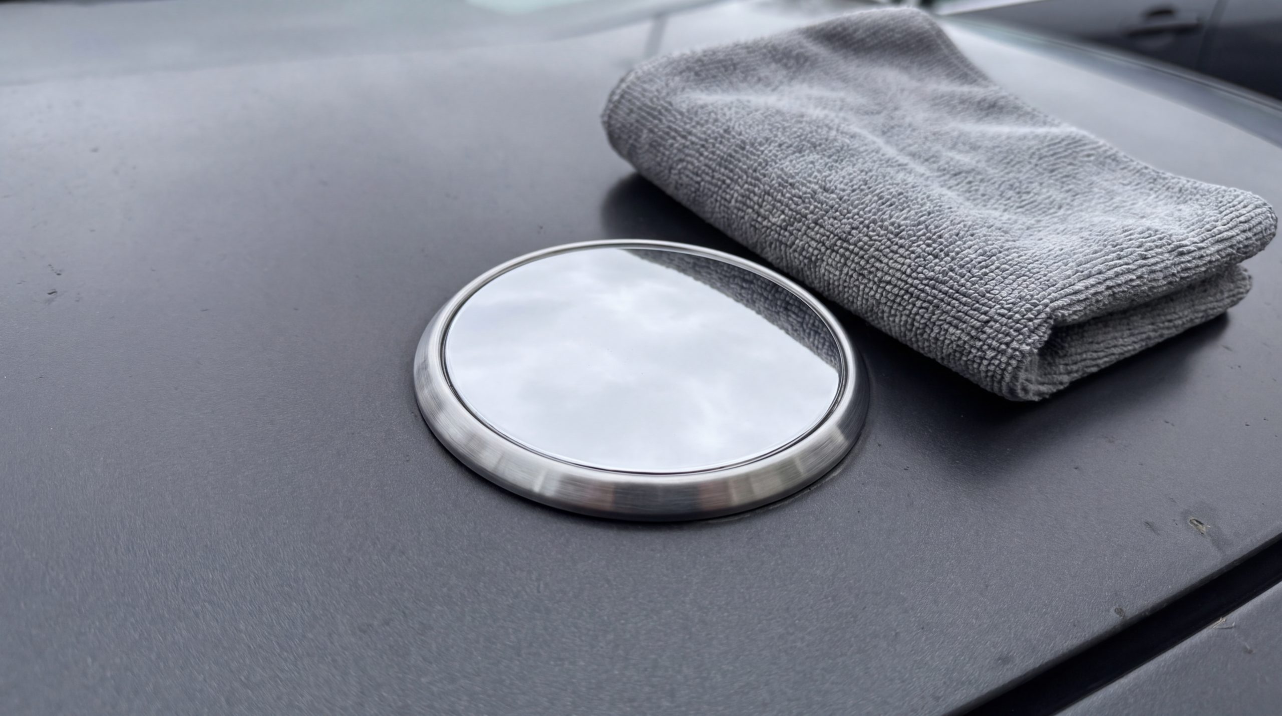 Ett tomt, runt kromemblem utan logotyp på en mörk motorhuv bredvid en mikrofiberduk